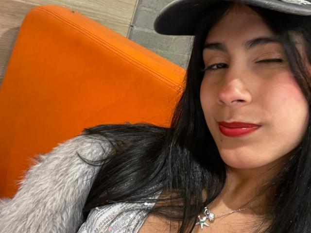 ValeryTranss hat eine mittelbraune Haut, schwarzes Haar, roten Lippenstift und eine Kreuzkette um den Hals, zwinkert leicht und lächelt dabei, und trägt eine graue Felljacke und eine schwarze Mütze..