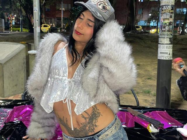 ValeryTranss with dark hair, wearing a gray cap, silver sequin top, furry jacket, showing a large tattoo on her right side, and blue jeans, standing outdoors at night.
