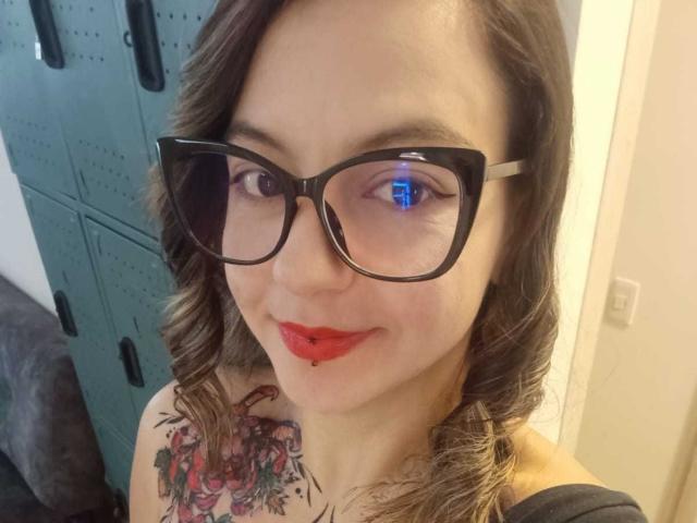 ArianaBlonde with glasses, red lipstick, and a tattoo on her shoulder, smiling at the camera, showing her tits slightly. Her blue eye is visible.