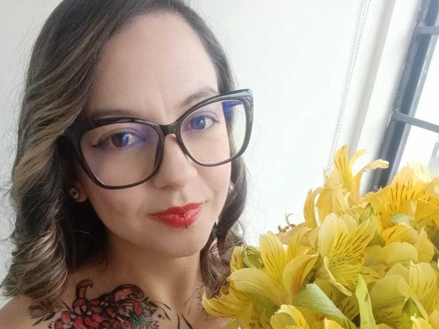 ArianaBlonde with glasses, red lipstick, curly brown hair, yellow lilies, and a red and black tattoo on her right shoulder, looking at the camera with blue eyes.