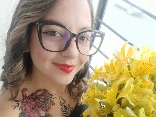 ArianaBlonde with glasses, red lipstick, and curly brown hair, smiling at the camera. She has a colorful tattoo of a red crab on her right shoulder. Yellow flowers.
