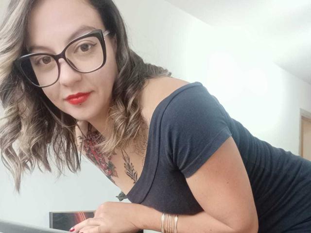 ArianaBlonde with glasses, red lipstick, and curly brown hair, wearing a black off-shoulder top, showing a tattoo on her chest, leaning forward with a confident smile.