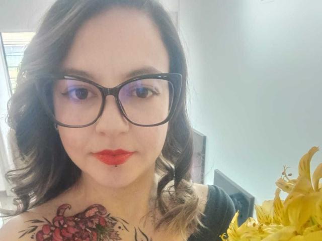 ArianaBlonde with red lips, black glasses, curly brown hair, and a colorful tattoo on her right shoulder, holding yellow lilies. Her tits are partially visible.