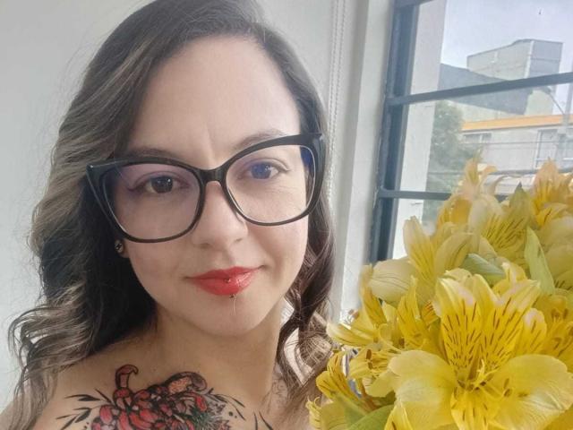 ArianaBlonde with wavy brown hair, black glasses, red lipstick, and a colorful tattoo on her shoulder, holding yellow lilies, smiling softly.