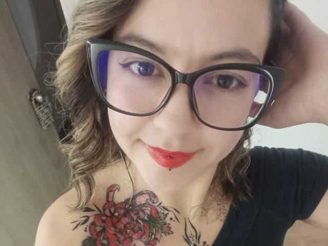 ArianaBlonde with glasses, red lipstick, and a lip piercing, has curly brown hair, black shirt, and a colorful tattoo on her left boob, smiling at the camera.