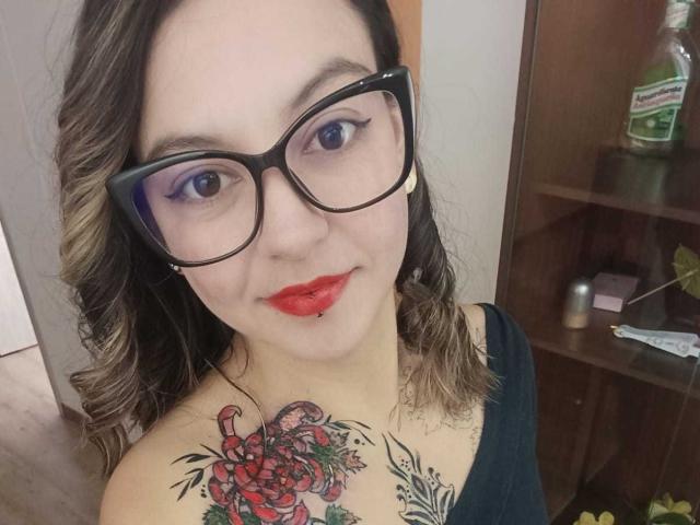 ArianaBlonde with glasses, red lipstick, and a nose ring, has curly brown hair and a large colorful tattoo of roses and a crab on her chest.