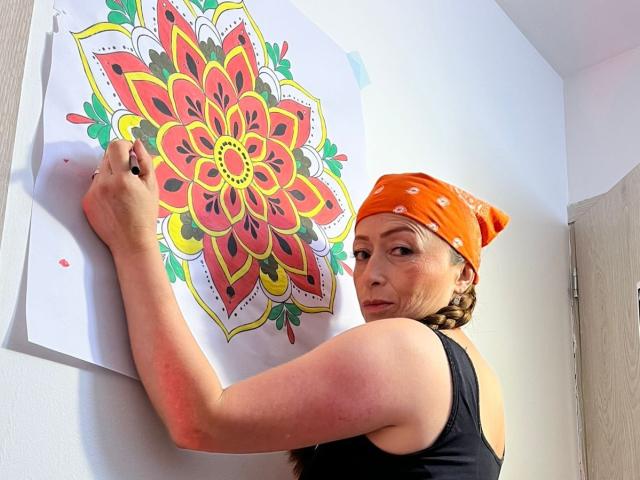 GianineLarose with orange bandana, black tank top, braided hair, painting colorful mandala on white wall, looking over shoulder, holding black marker, white room, door in background.