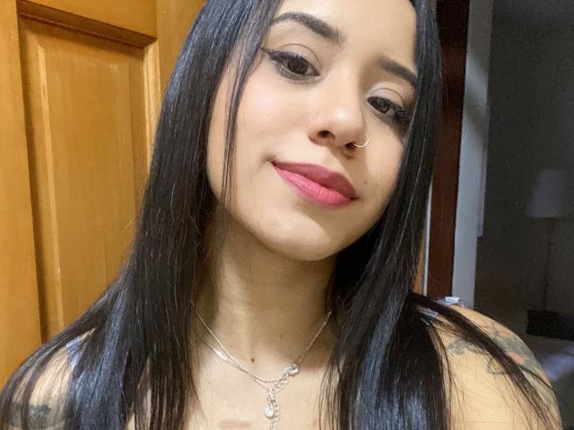 AsheleyMayer has long black hair, a nose ring, and pink lipstick. She's smiling with a slight tilt, wearing a necklace with a pendant, showing tattoos on her shoulder.