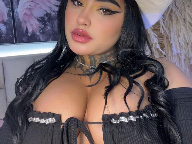 LairaHearvy with large tits, black wavy hair, yellow eye makeup, pink lips, black off-shoulder top, tattooed neck, and a white headscarf. Her cleavage is prominent.