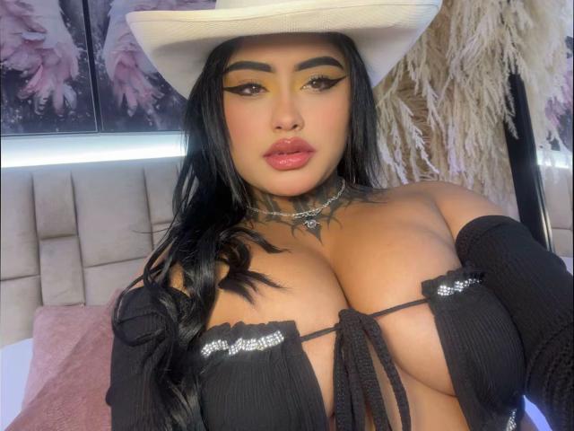 LairaHearvy with big tits, wearing a white cowboy hat, black off-shoulder top, and choker, has long black hair, bold eyeliner, and glossy pink lips, in a bedroom.