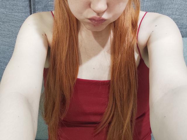 SheritaChieng tiene el pelo largo y rojo, labios carnosos y lleva una camiseta sin mangas roja. Sus brazos pálidos están levantados y está tomando una selfie. Se pueden ver los senos debajo de la camiseta..