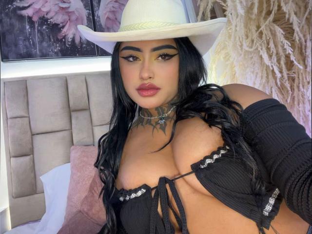 LairaHearvy with black hair, wearing a white cowboy hat, black off-shoulder top showing her big tits, yellow eyeliner, and a tattooed neck, in a bedroom setting.
