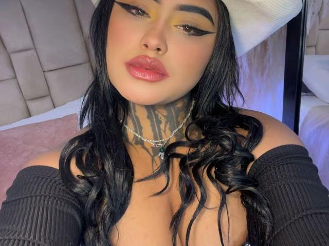 LairaHearvy with black wavy hair, thick eyeliner, pink glossy lips, necklace, and off-shoulder black top, showing tattoos on her neck. Boobs slightly visible, wearing a white hat.