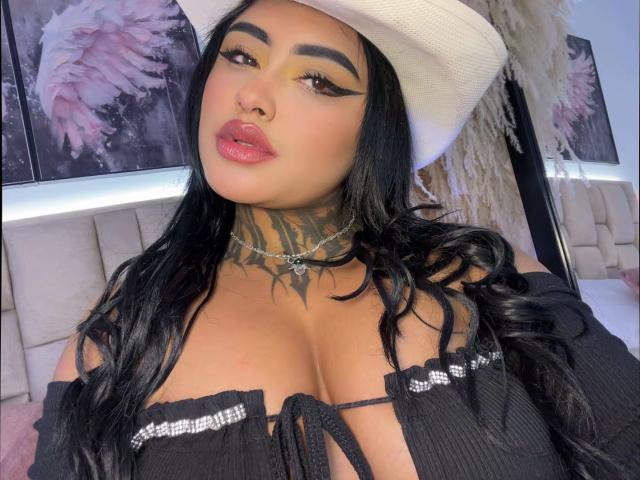 LairaHearvy with black hair, wearing a white cowboy hat, black off-shoulder top, showing her large tits, tattooed neck, and pink lipstick. Her makeup includes bold, winged eyeliner.