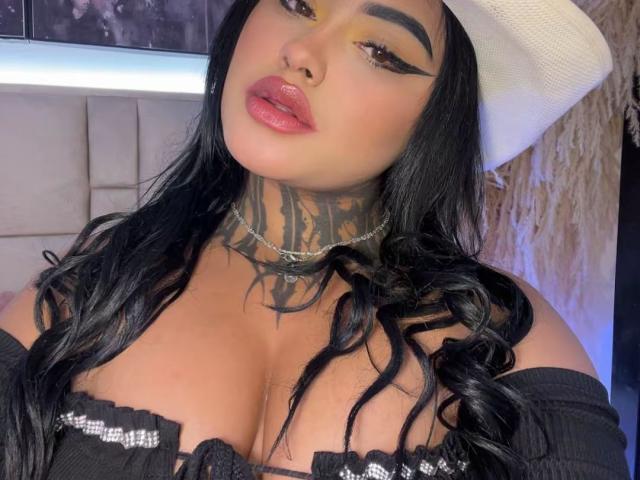 LairaHearvy with black wavy hair, white cowboy hat, thick black eyeliner, pink lips, tattoos on her neck, choker, and off-shoulder black top showing her big tits.