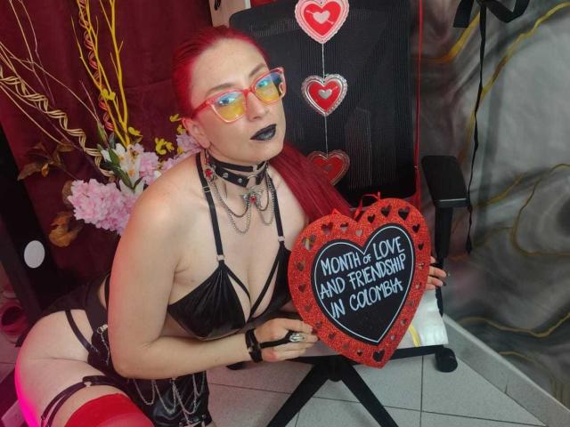 QueenScarletSecret met rood haar, zwarte lipstick, een bril, en zwarte, open lingerie, die een hartvorm maakt, draagt een choker en kettingen, en knielt naast een zwarte stoel..