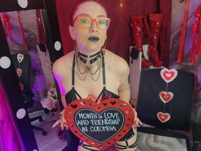 QueenScarletSecret met rode bril, zwarte lipstick, een zwart topje met bandjes en een hartje met de tekst "Mama en vriendschap in Coombr". Rode laarzen en reflecterende lampen op de achtergrond..