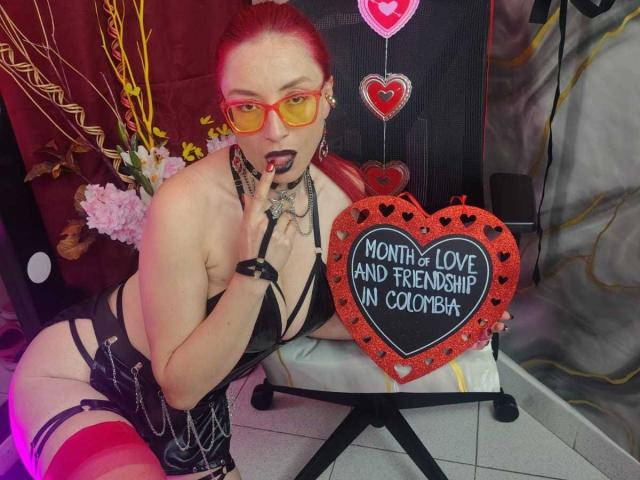QueenScarletSecret met rood haar, gele brillen, zwarte lingerie en rode kniekousen, die een hartvorm maakt, haar vinger zuigt, met zichtbare borsten en een zwarte choker..