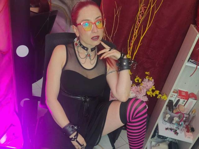 QueenScarletSecret avec des cheveux roux, des lunettes, une robe en maille noire, un collier, des manchettes, des bas roses et noirs à rayures, assise sur une chaise, la main près de la bouche, regardant vers le haut..