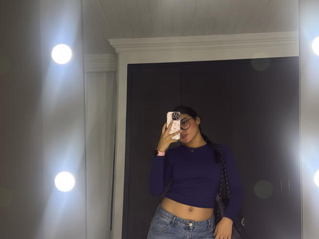 MiiaPalmer with dark skin, wearing glasses, a navy blue long-sleeve crop top, and blue jeans, taking a mirror selfie, showing her belly button, with a black braid.