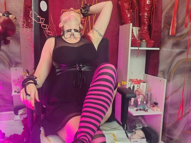 QueenScarletSecret con gli occhiali, vestito nero, cintura in pelle nera, calze rosa a righe che arrivano all'altezza coscia, polsini neri, braccio sollevato, seduta su una sedia, stivali rossi sullo sfondo, tatuaggi visibili..