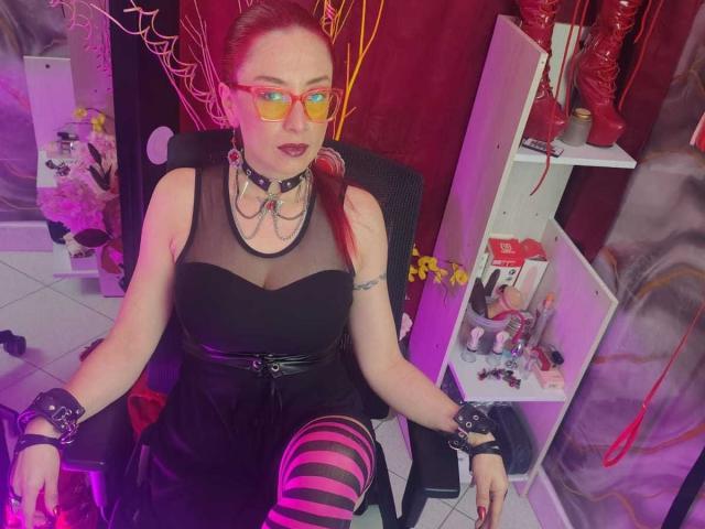 QueenScarletSecret met rode brillen, een zwarte kanten top, een zwarte leren rok, roze en zwarte streepmotieven op de kuiten, en zwarte polsbanden, zittend op een stoel, waarbij haar borsten zichtbaar zijn..