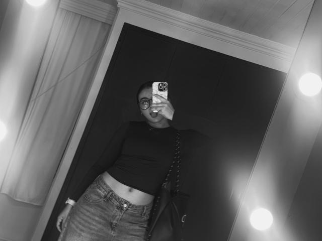 MiiaPalmer with glasses, black crop top, and high-waisted jeans, holding phone, showing her belly, mirror with lights, bag on shoulder, black background, mirror selfie, monochrome.