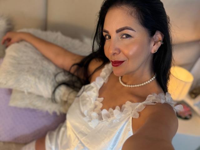 AlizonMoreti with long black hair, red lipstick, pearl necklace, white frilled dress, smiling, lying on a bed, arm raised, visible boobs, soft lighting, warm bedroom, relaxed vibe.