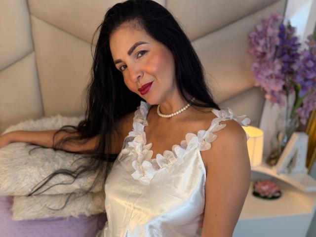 AlizonMoreti with long black hair, wearing a white dress with ruffle straps, pearl necklace, red lipstick, smiling, resting on a white pillow, purple flowers in the background.