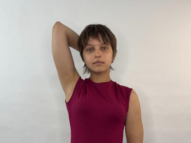 Alia Kalafut, com cabelo castanho curto, vestindo uma blusa vermelha sem mangas, braço levantado, olhando diretamente para frente, mostrando seios pequenos, um leve volume nas nádegas e uma expressão neutra..