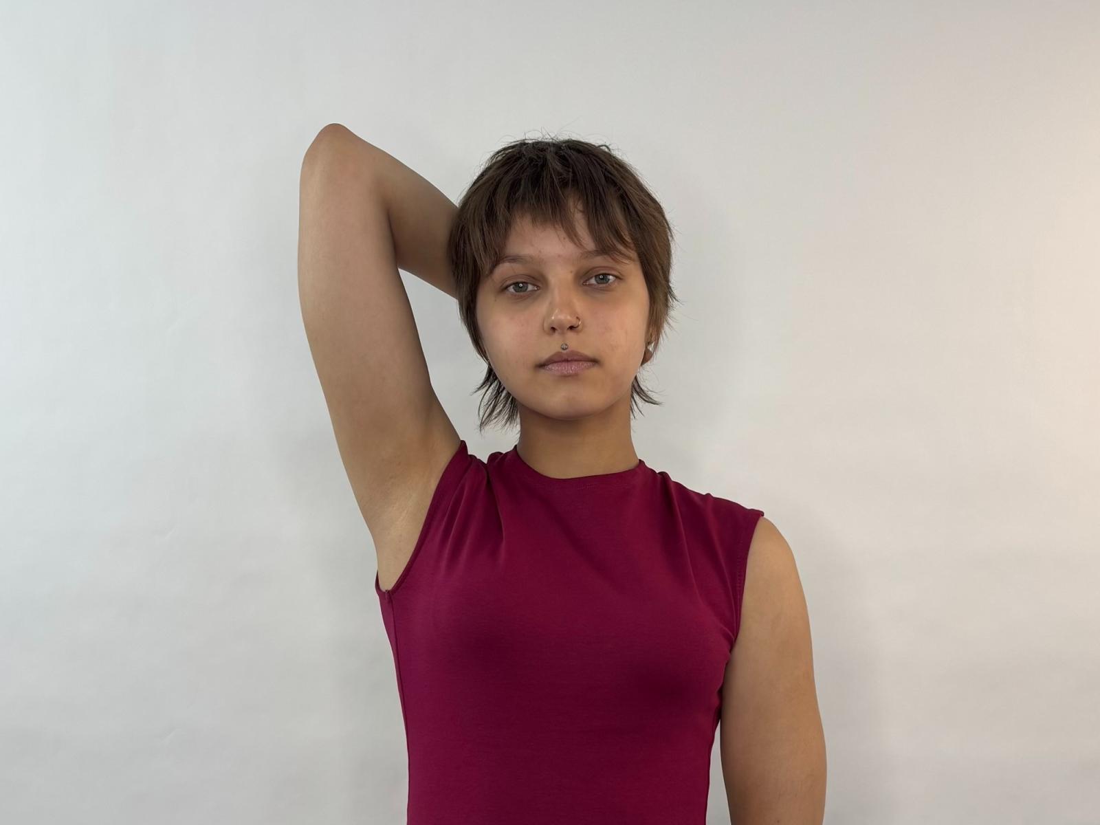 AliaKalafut with short brown hair, wearing a sleeveless red top, arm raised, looking straight ahead, showing small tits, slight ass, and a neutral expression.