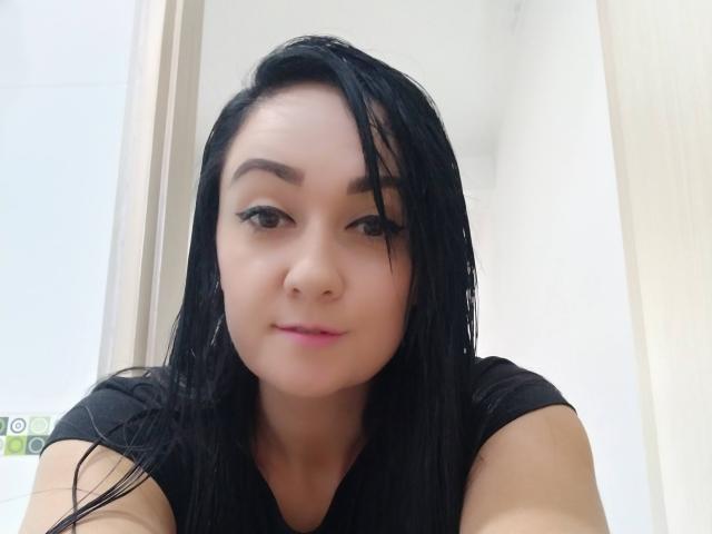 Katnnis has wet black hair, brown eyes, and pink lipstick, wearing a black shirt, looking at the camera with a slight smile, in a bright room.