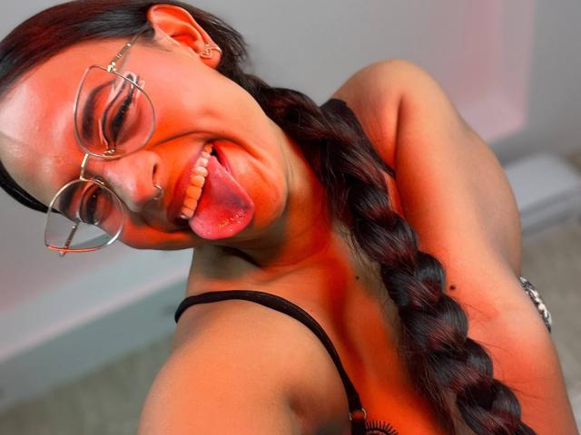 LilithRussoo with glasses, nose ring, and braided red hair, sticking out her tongue, showing her teeth, in a selfie with red light. Boobs visible in black top.