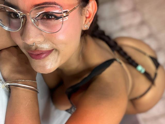 LilithRussoo with glasses, nose ring, and earrings, lying down in black lingerie, showing her ass and small tits, smiling with a slight blush, braided hair, silver bracelets.