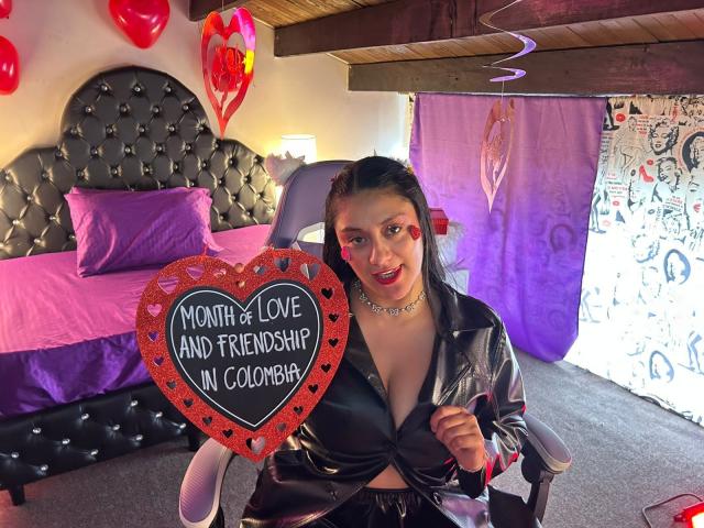 CristalZafiro with dark hair, red lipstick, and a black leather jacket, holding a heart-shaped sign saying "Month of Love and Friendship in Colombia." Boobs visible under jacket.