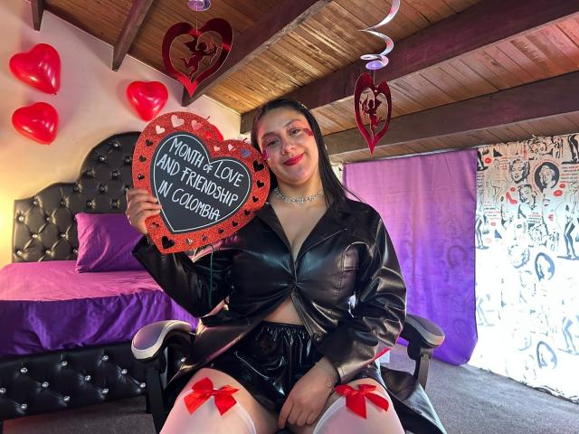 CristalZafiro in black leather outfit, holding heart-shaped sign, red bows on white stockings, purple bed, wood ceiling, smiling, long black hair, makeup, wood ceiling, Christmas decor.