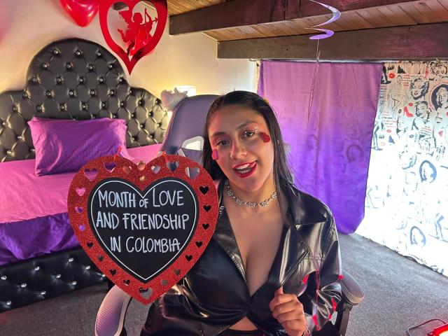 CristalZafiro with dark hair, red heart makeup, black shiny jacket, showing heart sign with "Month of Love and Friendship in Colombia", large tits, smiling, in colorful room.