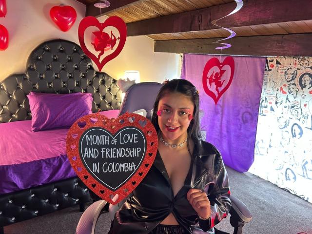 CristalZafiro with tits visible in a black leather jacket, holding a heart sign, smiling in a room with pink bedding, red hearts, and a purple curtain.