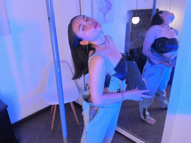 Iniridax with long black hair, wearing blue denim overalls, black strapless top, and white gloves, looking up, reflection in mirror, blue light, standing by pole in modern room.