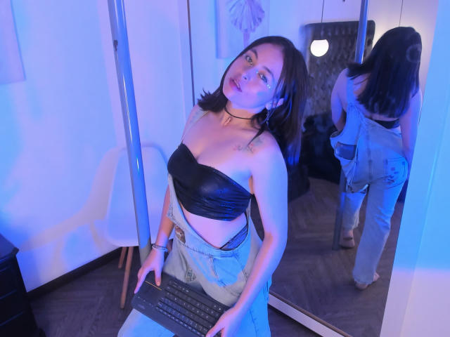 Iniridax with black strapless top, showing small tits, wearing blue jeans, standing by a mirror, holding a laptop, in a blue-lit room, reflected in the mirror.