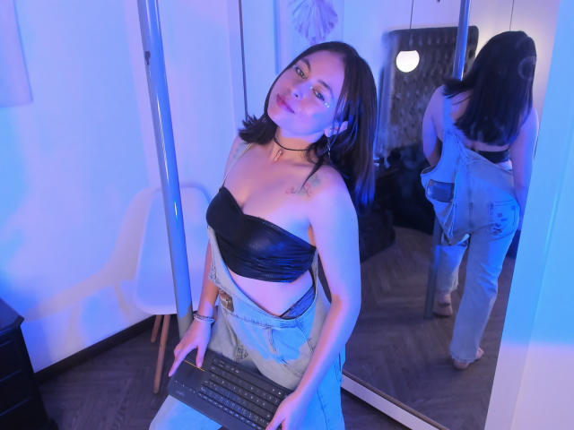 Iniridax with brown hair, black strapless top, showing tits, and blue jeans, sits at a laptop, reflected in a mirror, wearing a choker, in a blue-lit room.