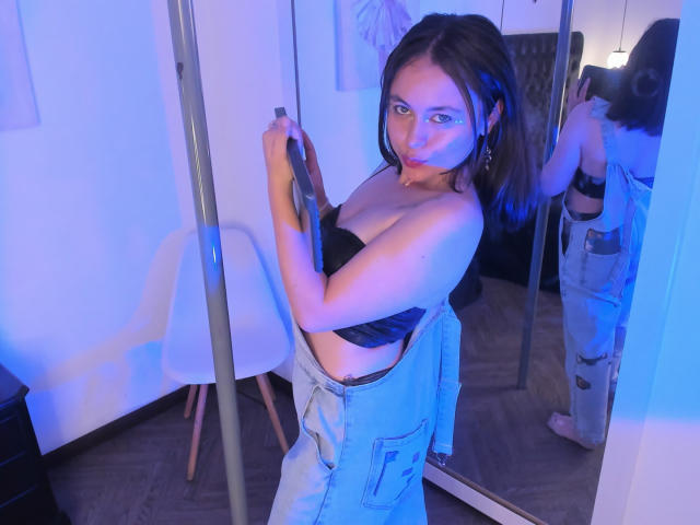 Iniridax with medium tits, black strapless top, blue jeans, holding a pole, reflected in mirror, blue lighting, brown hair, earrings, standing in a room with a chair.