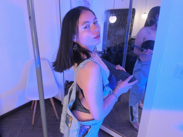 Iniridax with dark hair, blue eyes, wearing a denim jacket, black bra, and jeans, holding a game controller, reflected in a mirror, showing her ass. Blue room lighting.