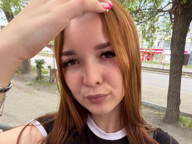 HottiePurdum with reddish-brown hair, slightly pouty lips, and a cute face, touching her head with a hand, showing pink nails, wearing a black and white top.
