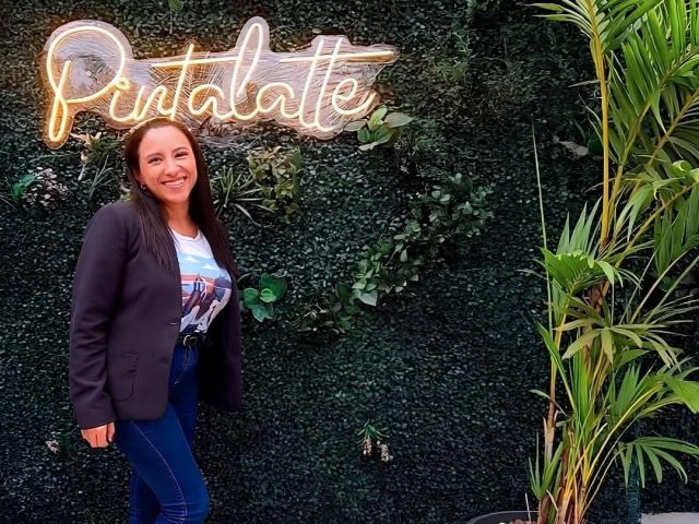 Jade Foster se tient debout, souriante, vêtue d'un blazer noir, d'un jean bleu et d'un t-shirt blanc avec un motif, avec un panneau néon "Pantallette" au-dessus d'elle et du feuillage vert sur la droite..