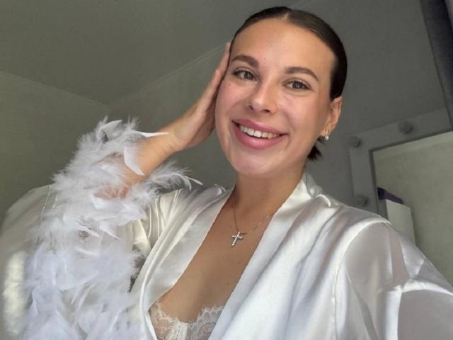 BlackFatal with brown hair, smiling, wearing a white satin robe with feather trim, lace bra, and cross necklace, hand on cheek, showing white teeth, and cute earrings.