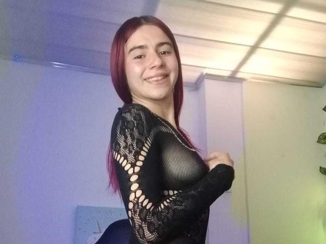 LoveMaxim with red hair, smiling, wearing a black mesh top showing her small tits, holding her top. Indoor lighting, ceiling tiles, and a plant in the background.
