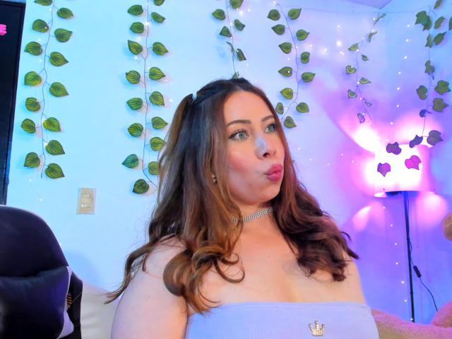 JennyLawry with puffy lips, brown wavy hair, wearing a choker and strapless top, showing her boobs, in a room with purple and blue lights, and hanging vines.
