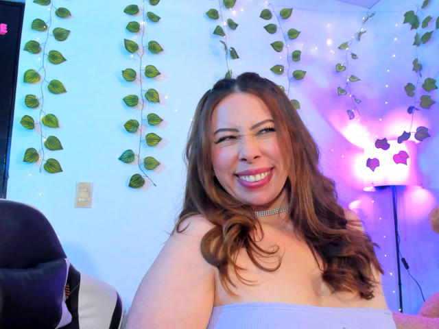 JennyLawry with wavy brown hair, smiling, wearing a strapless purple top, choker, and pink lipstick, showing her tits, in a room with green leaf garlands and purple lights.