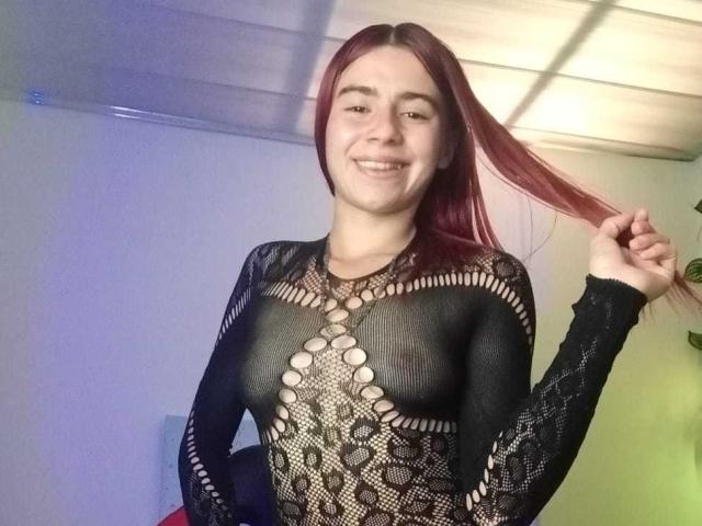 LoveMaxim met rood haar, lachend, gekleed in een zwarte kanten top die haar kleine, stevige borsten en tepels onthult, haar haar vasthoudend, in een heldere, binnenruimte..
